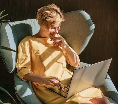 A smiling woman in a yellow dress uses a laptop while sitting in a blue armchair.